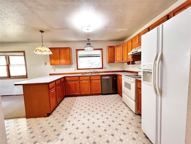 Kitchen featuring white appliances, a peninsula, brown cabinetry, a textured ceiling, and hanging light fixtures