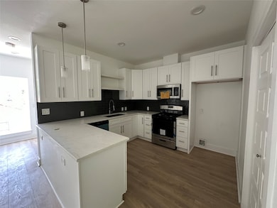 This kitchen features modern white cabinetry with sleek hardware, a black tile backsplash, and stainless steel appliances. The space includes a spacious island with a light countertop and pendant lighting, complemented by wood flooring for a contemporary look.