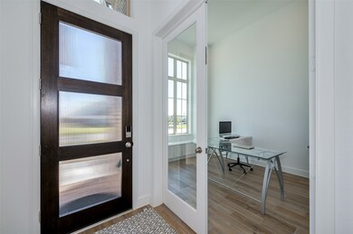 Modern entryway with a stylish frosted glass front door and a glass-paneled door leading to a bright office space. The office features  large windows allowing ample natural light. The flooring is a light wood-like ceramic tile, creating a warm and inviting atmosphere.