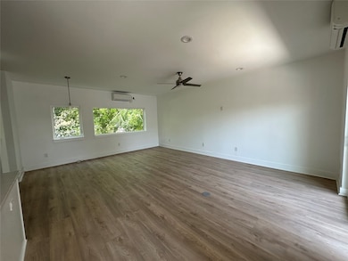 This spacious room features laminate wood flooring, large windows with a view of greenery, white walls, and modern lighting. It includes a ceiling fan and air conditioning for comfort.