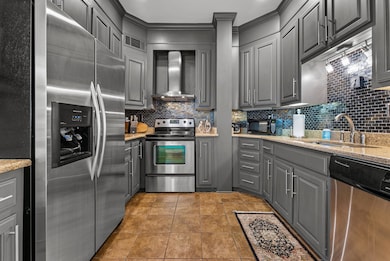 Kitchen featuring gray cabinetry, wall chimney exhaust hood, appliances with stainless steel finishes, sink, and light stone countertops.