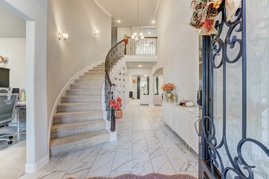 This elegant entryway features a grand curved staircase, marble flooring, and a welcoming chandelier. Decorated with seasonal accents, it leads to a cozy office space and a spacious living area, showcasing a blend of openness and sophistication.