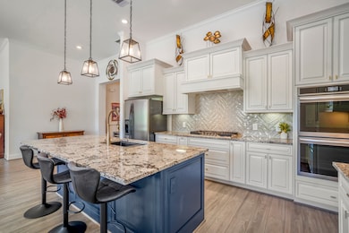 This kitchen features a spacious island with granite countertops and bar seating, pendant lighting, and stainless steel appliances. It boasts elegant white cabinetry, a herringbone tile backsplash, and ample natural light, creating a modern and inviting atmosphere.