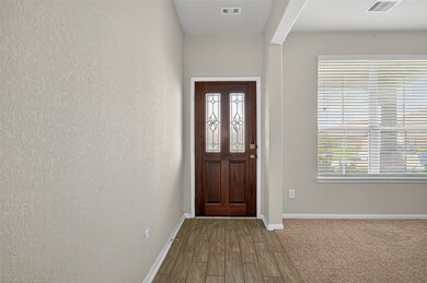 The photo shows a welcoming entryway with a stylish wooden front door featuring decorative glass panels. The space has a mix of wood-look flooring and carpet, with a large window allowing natural light to brighten the area.