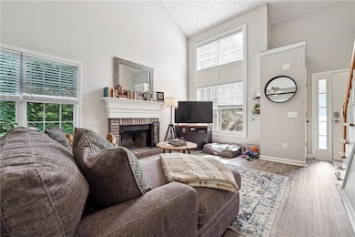 Living area featuring high vaulted ceiling, healthy amount of natural light, wood finished floors, a brick fireplace, and baseboards