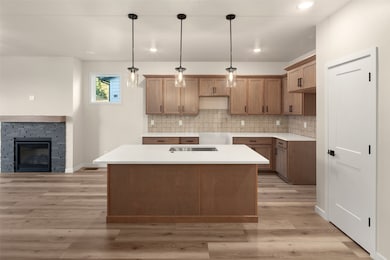 Kitchen with brown cabinetry, backsplash, decorative light fixtures, a center island with sink, and light stone counters
