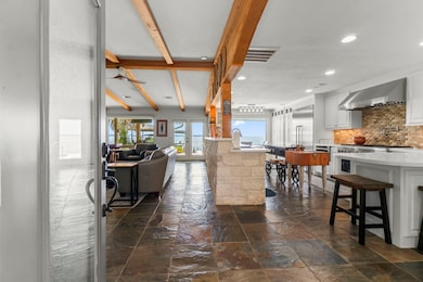 This open-concept home features a spacious kitchen with modern appliances and a stone backsplash, leading into a cozy living area with exposed wooden beams and slate flooring. Large windows offer plenty of natural light and lake views, creating a seamless indoor-outdoor feel.