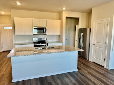 Kitchen with stainless steel appliances, light stone countertops, white cabinetry, dark wood-type flooring, and a center island with sink