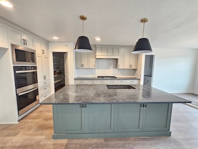 Kitchen with backsplash, stainless steel appliances, dark stone countertops, decorative light fixtures, and a center island with sink