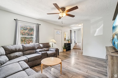 Living area with a textured ceiling, light wood-type flooring, ceiling fan, and stairway
