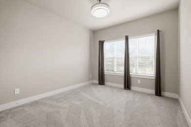 First floor: Secondary bedroom ideal for a guest, nursery or multigenerational use. This photo shows a bright, neutral-toned room with carpeted flooring. It features a large window with blinds and curtains, allowing natural light to fill the space. The ceiling has a modern light fixture, creating a clean and inviting atmosphere.