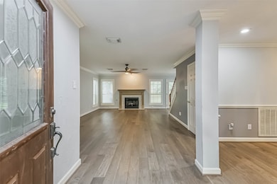 Welcoming entryway leading into a spacious living area with hardwood floors and a fireplace. The room is bright, featuring large windows with shutters, and there's a staircase visible on the right. The neutral color palette and crown molding add a touch of elegance.