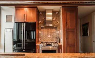 Kitchen with brown cabinetry, stainless steel gas range, wall chimney range hood, high end black refrigerator, and tasteful backsplash