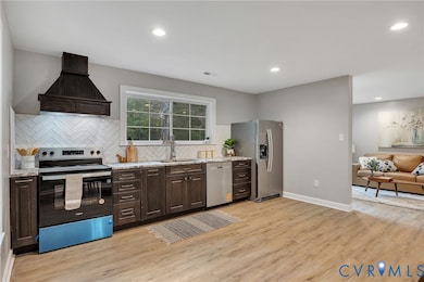 Kitchen with appliances with stainless steel finishes, dark brown cabinetry, custom range hood, tasteful backsplash, and light wood-type flooring