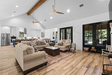 This spacious living area features a vaulted ceiling with exposed beams, modern ceiling fans, and large windows offering plenty of natural light. The open layout seamlessly connects to a stylish kitchen with high-end appliances, while the warm wood flooring and cozy seating area create an inviting atmosphere.