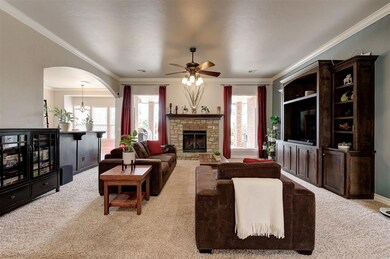 Carpeted living area with visible vents, arched walkways, ceiling fan, a stone fireplace, and ornamental molding