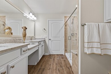 Ensuite Primary bathroom features a double vanity with granite countertops, center vanity area, extra cabinetry over the toilet, a large mirror, elegant lighting, and a glass-enclosed shower with tile accents. The wood-look floor adds warmth, and modern fixtures enhance the overall aesthetic.