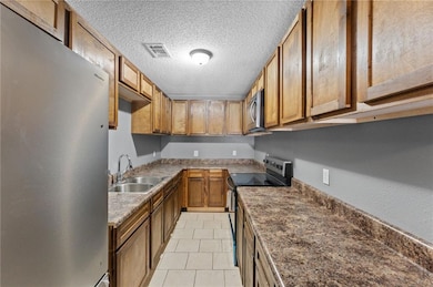 Kitchen featuring appliances with stainless steel finishes, a textured ceiling, light tile patterned flooring, brown cabinetry, and dark countertops