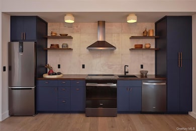 Kitchen featuring open shelves, blue cabinets, stainless steel appliances, wall chimney exhaust hood, and light wood-style floors