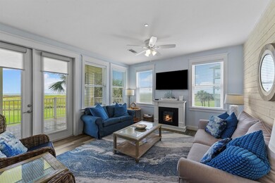 The open living room is filled with natural light from walls of windows and showcases a cozy mock fireplace, new flooring, and a breezy Gulf-inspired ambiance.