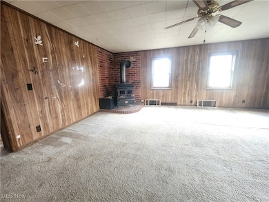 Unfurnished living room with a wood stove, carpet floors, wood walls, and a ceiling fan