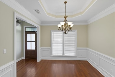 Unfurnished dining area with a tray ceiling, a chandelier, a wainscoted wall, crown molding, and dark wood-type flooring
