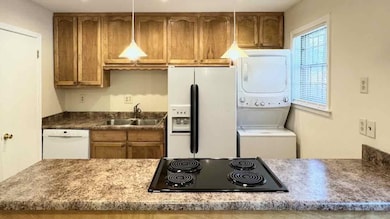 Kitchen with brown cabinetry, white appliances, stacked washer / dryer, dark countertops, and pendant lighting