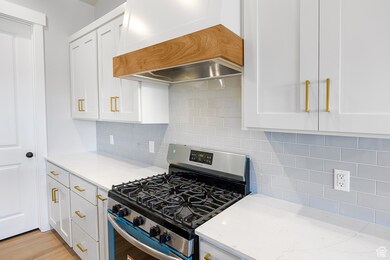 Kitchen featuring stainless steel range with gas cooktop, custom range hood, backsplash, white cabinetry, and light stone counters