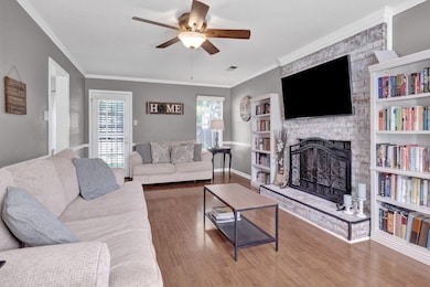Living area with a ceiling fan, a stone fireplace, wood finished floors, crown molding, and baseboards