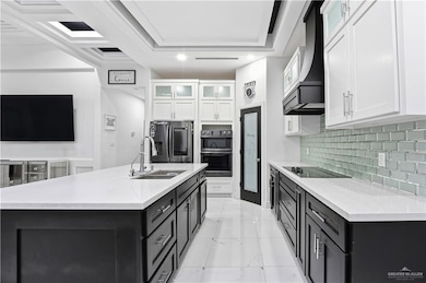 Kitchen with dark cabinetry, glass insert cabinets, stainless steel fridge, white cabinetry, and light stone counters