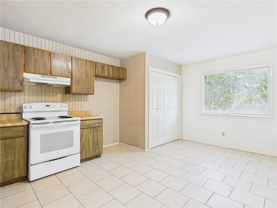 Kitchen featuring electric range, light countertops, under cabinet range hood, brown cabinetry, and light tile patterned flooring