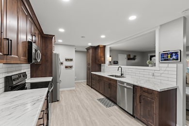 Kitchen featuring decorative backsplash, stainless steel appliances, light stone countertops, dark brown cabinets, and light wood-style flooring
