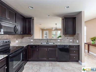The kitchen looks out to the great room. The black, brown, and cream colored granite counters unite the deep toned cabinets and black appliances to the light tiled floors and decorative back-splash.