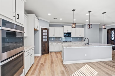 Kitchen featuring stainless steel appliances, decorative light fixtures, light stone counters, a large center island with sink, and white cabinetry.
