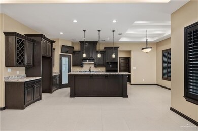 Kitchen with dark brown cabinets, light stone countertops, a raised ceiling, an island with sink, and decorative light fixtures