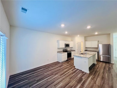 Kitchen with dark hardwood / wood-style floors, stainless steel appliances, white cabinets, light stone countertops, and a kitchen island with sink
