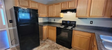 Kitchen featuring black appliances, under cabinet range hood, dark countertops, and light brown cabinetry