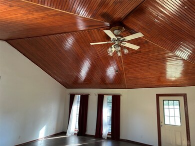 This room features a high, varnished beadboard ceiling with a central fan and light fixture. It has large windows with curtains, providing natural light, and a door with glass panels. The walls are light, creating a spacious and airy feel.