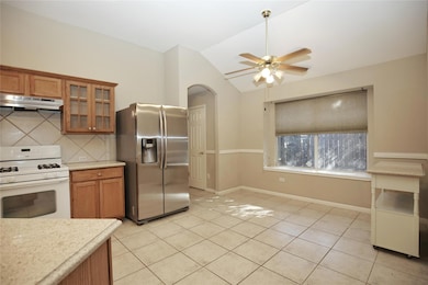Kitchen with white gas stove, stainless steel fridge with ice dispenser, brown cabinetry, light tile patterned floors, and vaulted ceiling