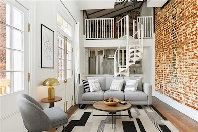 Sunlit living room with exposed brick, original hardwood floors, and French doors opening to the private balcony. Virtually staged using artificial intelligence.