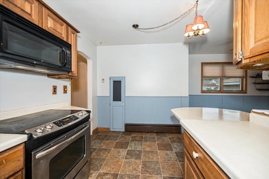 Kitchen with stainless steel electric range, black microwave, brown cabinetry, light countertops, and a wainscoted wall