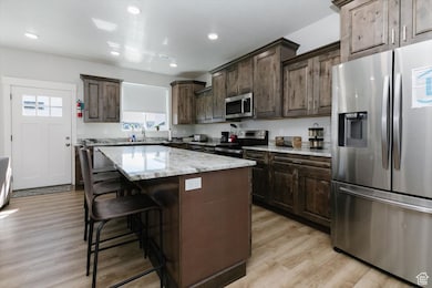 Kitchen with appliances with stainless steel finishes, a breakfast bar area, dark brown cabinetry, light stone countertops, and recessed lighting