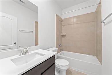 This first floor secondary bathroom offers tile flooring, a bath/shower combination with a stylish tile surround, rich stained wood cabinetry, elegant light countertops, a mirror, and sleek fixtures with modern finishes.