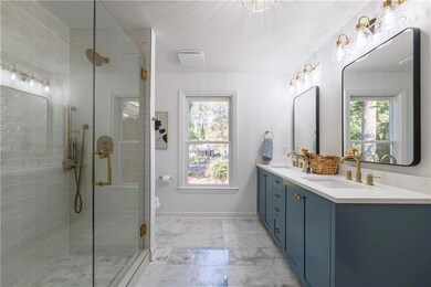 The luxurious primary bathroom showcases ocean white marble floors, double sink vanity, and a shower with both rain and standard shower heads, accented with pearl-textured subway tile and sophisticated black and brass fixtures.