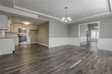 Unfurnished living room featuring a tray ceiling, a chandelier, dark wood-style flooring, a ceiling fan, and wainscoting
