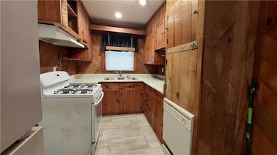 Kitchen with white appliances, light countertops, brown cabinetry, under cabinet range hood, and wooden walls