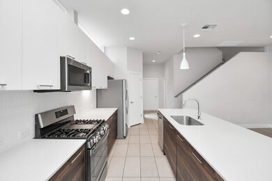 Modern kitchen with stainless steel appliances, white countertops, dark cabinetry, and a staircase to the upper level. Bright and clean with pendant lighting.