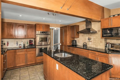 Kitchen featuring black appliances, backsplash, wall chimney exhaust hood, dark stone countertops, and brown cabinetry