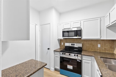 Kitchen with stainless steel appliances, decorative backsplash, white cabinets, light wood-style flooring, and dark stone countertops