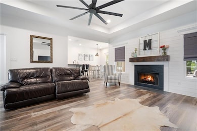 Living room with a fireplace with flush hearth, wood finished floors, a raised ceiling, a ceiling fan, and recessed lighting